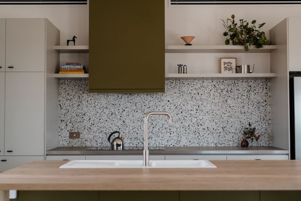 A contemporary kitchen viewed straight on, with pale built-in cabinetry, open display shelving, a large olive-green rangehood canopy, a speckled terrazzo-style splashback, stainless steel tap and kettle, and a broad light timber island with an inset double sink. The restrained styling includes books, ceramics, framed artwork and plants on the shelves.