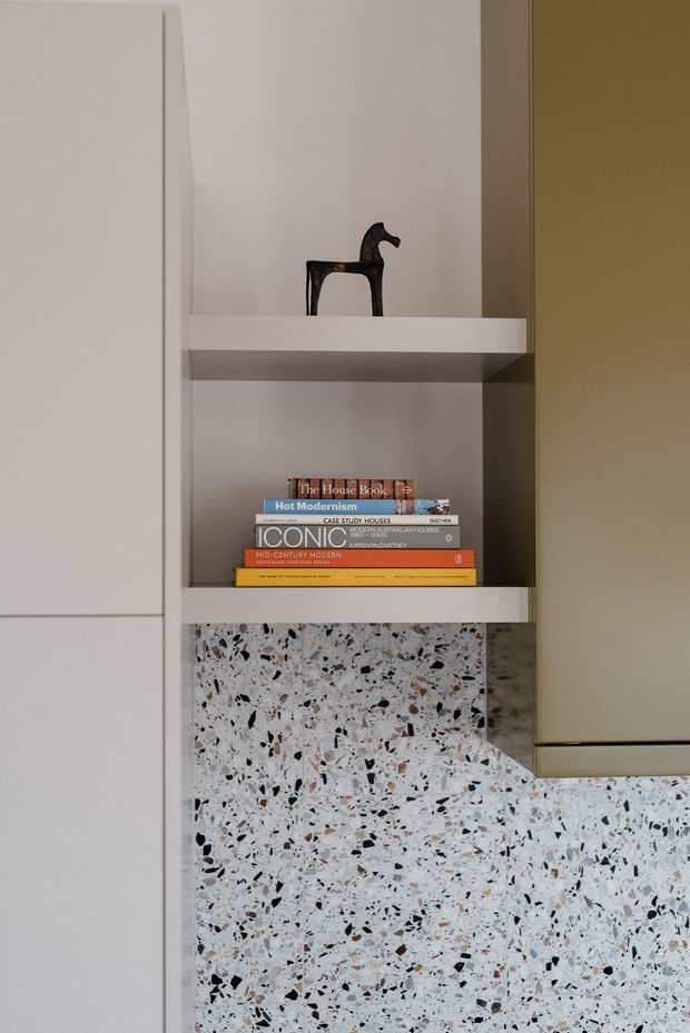 Close-up view of a modern built-in shelving and cabinetry detail. White open shelves display a small dark horse sculpture and a stack of design books, while a pale terrazzo backsplash or wall surface with black and warm aggregate flecks continues below. A muted olive-grey cabinet panel is visible at right, creating a restrained, contemporary material palette.