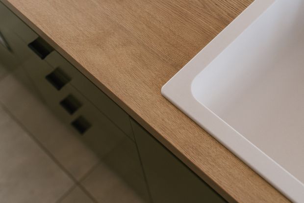 Close-up detail of a contemporary bathroom vanity, showing a light timber-look benchtop with an integrated or undermount white rectangular basin and dark green fixed cabinetry with black hardware. Large-format neutral floor tiles are visible below.
