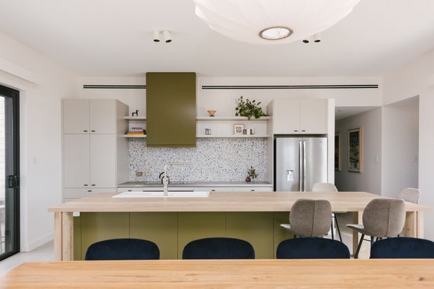 Bright contemporary open-plan kitchen and dining area with a large pale timber island, olive-green island cabinetry and rangehood, white cabinetry, stainless-steel refrigerator, and a terrazzo-style speckled backsplash. Dark upholstered stools and neutral dining chairs surround the island, while a sculptural white ceiling light adds a soft focal point.