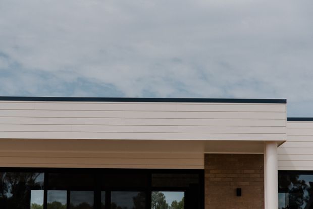A close-up exterior view of a contemporary, low-profile building facade under an overcast sky. The composition shows horizontal pale cladding, a dark flat roof edge, a deep covered entry or terrace, large dark-framed glazing, a warm tan brick feature wall, and a white structural column. Trees are faintly reflected in the glazing.