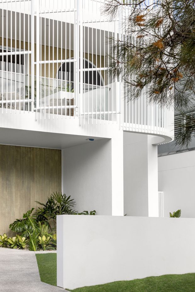 Exterior view of a contemporary white multi-level residence, featuring curved balcony edges, vertical white metal screening, white rendered walls, large glazed openings, and a timber-clad entry or garage door. Tropical planting, a low white boundary wall, lawn, and a concrete walkway frame the facade.