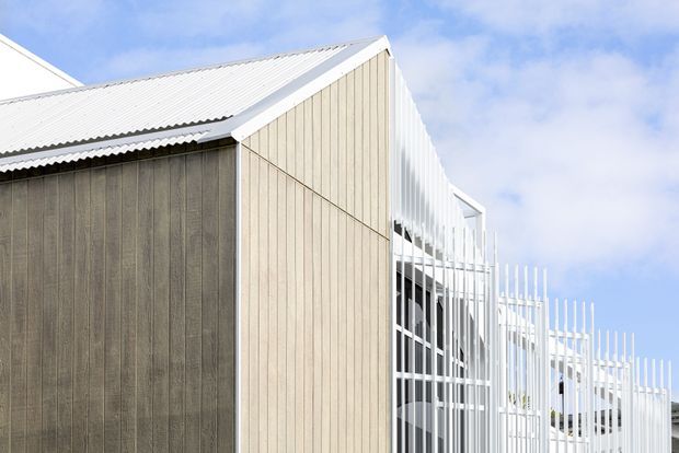 Exterior architectural view of a contemporary building with a prominent pitched corrugated roof, pale vertical timber-like cladding, and a striking white vertical slatted screen over glazed facade areas. The composition is set against a bright blue sky and focuses on the building envelope and roof form.