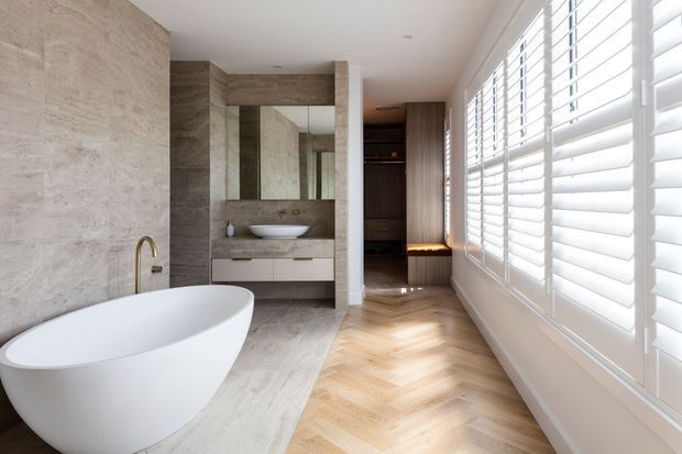 A bright, refined contemporary bathroom featuring a freestanding white oval bathtub, wall-mounted double vanity with vessel basin, large mirror, and warm brass tapware. The room is finished with large-format beige stone-look wall and floor surfaces, timber herringbone flooring, dark timber built-in cabinetry, and expansive white plantation shutters along the right wall.