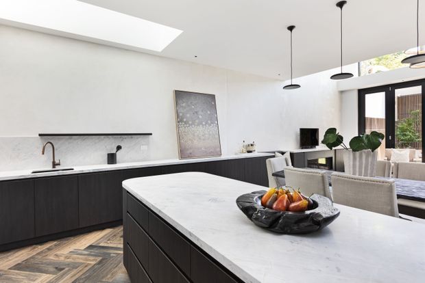 Bright contemporary open-plan kitchen and dining area with a large pale stone island, dark linear cabinetry, integrated sink and tap, minimalist black pendant lights, and a skylight. Herringbone timber flooring adds warmth, while glazed doors connect to an outdoor area. A dining table and chairs are visible beyond the island.