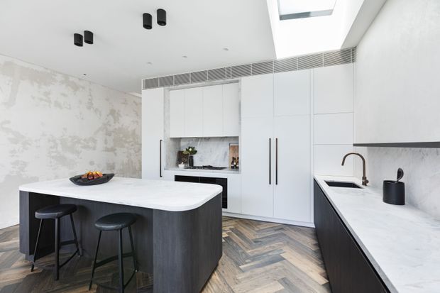 Bright contemporary kitchen with a large white waterfall island, dark vertical-grain cabinetry, integrated white wall storage, and a long perimeter benchtop with undermount sink. The room features a herringbone timber floor, white marble-look surfaces, black fixtures and hardware, a textured pale feature wall, ceiling ventilation grille, and skylight.