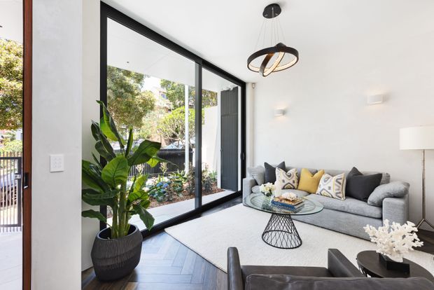Bright contemporary living room with a large grey sectional sofa, patterned and yellow cushions, glass coffee table, sculptural pendant light, and floor-to-ceiling black-framed glazing opening toward a planted courtyard. The room has white walls, dark timber flooring laid in a chevron pattern, and a restrained monochromatic palette with warm yellow accents.