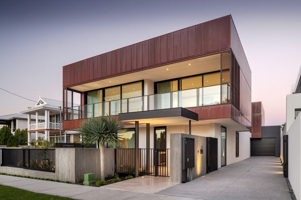 Contemporary two-storey residence with a strong rectilinear form, clad in dark reddish-brown vertical metal panels over a light rendered lower level. Large glazed openings, a projecting balcony with glass balustrade, recessed warm lighting, a covered entry, and an integrated single garage define the street-facing facade. A landscaped front garden sits behind a dark metal fence and concrete boundary wall.
