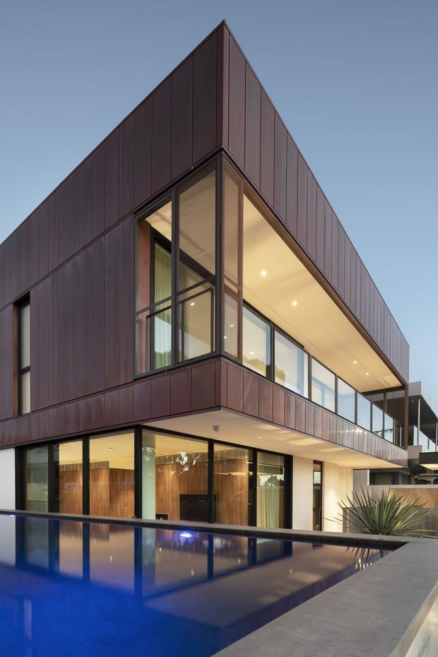 A contemporary multi-storey residence photographed at dusk, with a sharply folded gable-like roof form and dark reddish-brown vertical cladding. Extensive floor-to-ceiling glazing opens onto a covered upper-level terrace and overlooks a reflective swimming pool in the foreground. Warm interior lighting, clean cantilevered volumes, and dark metal-framed windows create a refined modern composition.