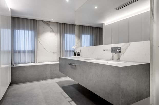 A spacious contemporary bathroom with a large freestanding-style bathtub beneath sheer curtains, a broad floating double vanity with integrated basins, wall-mounted taps, and a large mirrored cabinet. The room features grey stone-look surfaces, white marble-look wall panels with veining, recessed ceiling lighting, and a restrained monochromatic palette.