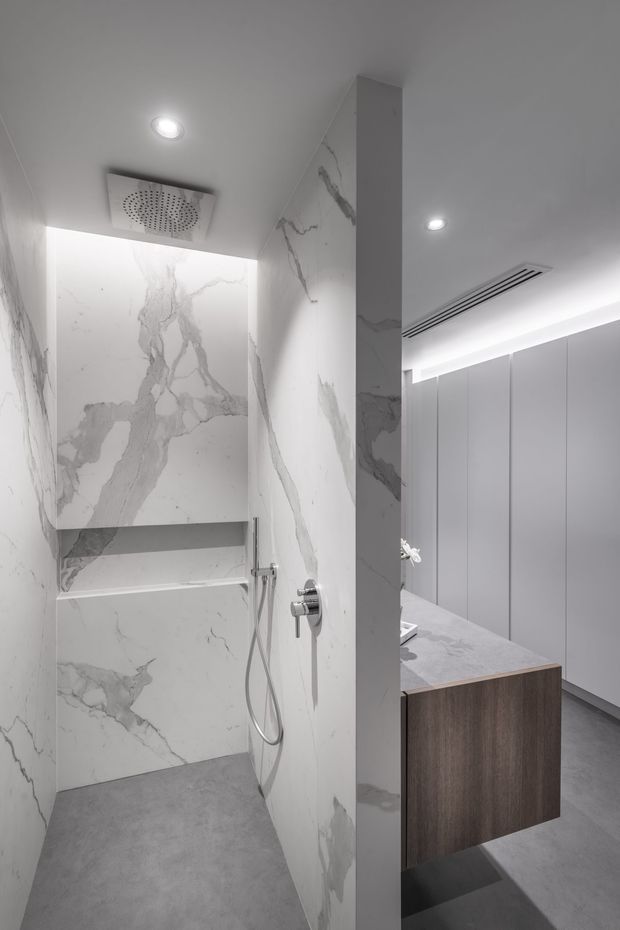 Contemporary bathroom featuring a walk-in shower clad in white marble-look slabs with pronounced grey veining, a concealed rain shower and handheld fitting, grey concrete-look flooring, and a freestanding timber-toned vanity with a pale stone benchtop. Full-height grey cabinetry and recessed linear ventilation add to the clean, minimalist fit-out.