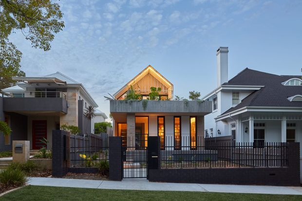 Street-facing view of a contemporary two-storey residence with a flat-roofed concrete lower volume, warm timber-clad gabled upper structure, extensive glazing, and a dark metal boundary fence. Neighbouring homes flank the property, and the scene is photographed at dusk with warm interior lighting visible.