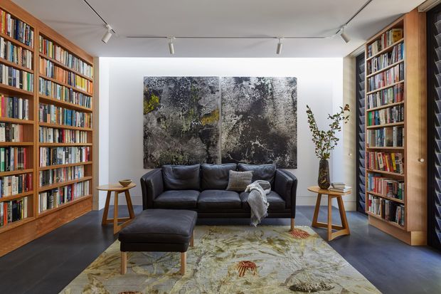 A refined home library and lounge with floor-to-ceiling timber bookshelves lining both side walls, a black leather sofa and ottoman, small timber side tables, and a large abstract artwork. Track lighting illuminates the white ceiling, while a large patterned rug softens the dark floor and creates a warm, sophisticated reading space.