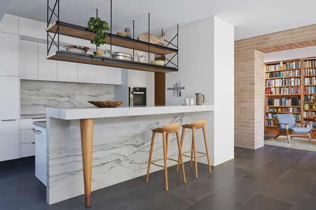 Bright open-plan kitchen with a large white veined stone island and breakfast bar, pale integrated cabinetry, timber bar stools, and black-framed open shelving with decorative objects and plants. A brick-lined opening leads to a book-lined living or library area with a mid-century-style lounge chair. Dark tiled flooring and white walls create a clean, contemporary interior.