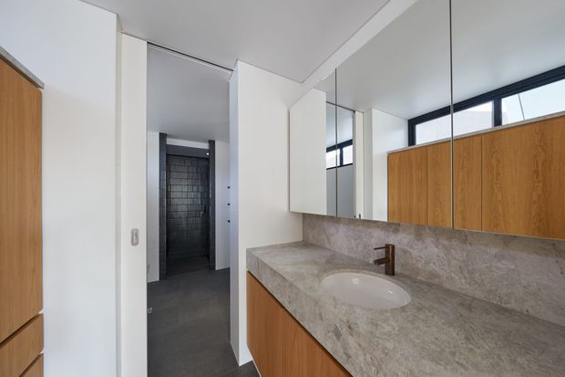 Contemporary bathroom vanity with a long stone benchtop, integrated white basin, timber cabinetry and bronze tapware. Full-height mirrors reflect the room, while high horizontal windows bring in natural light. A dark tiled shower area is visible through the doorway.