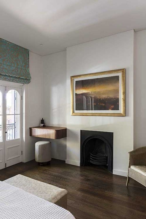 A refined living room with white walls, dark timber flooring, a black arched fireplace, framed artwork, and a compact wall-mounted timber console with a round upholstered stool. A patterned Roman blind and glazed French door provide architectural detail, while a textured sofa or bed-like seating element occupies the foreground.