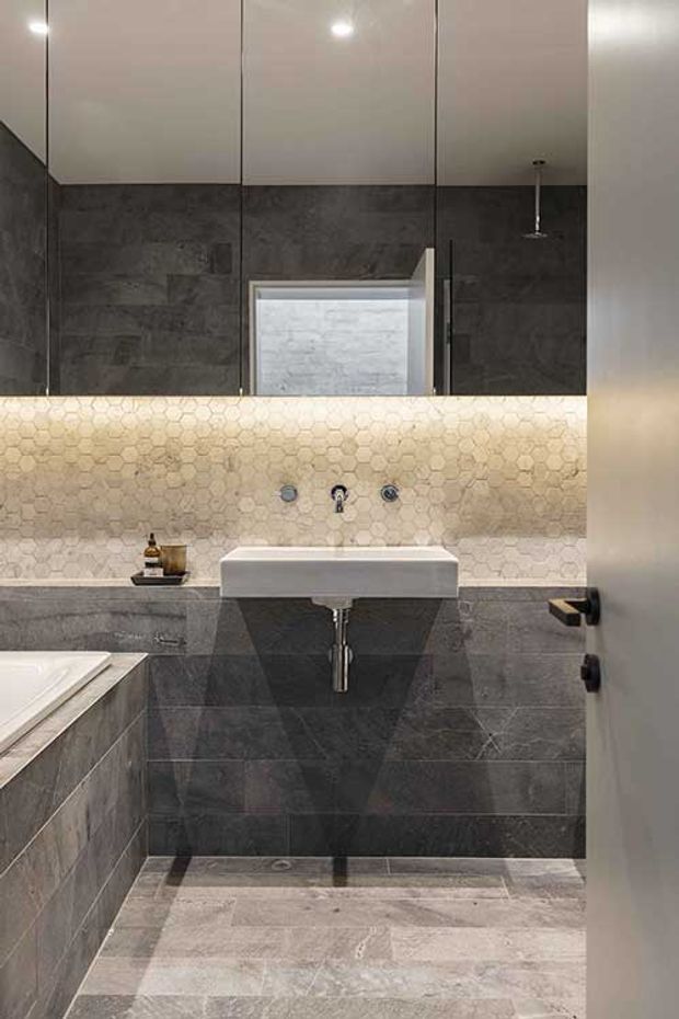 A compact contemporary bathroom viewed through a partially open door. The room features dark grey stone-look wall tiles, a light grey tiled floor, a wall-mounted white basin with exposed chrome plumbing, a large mirrored cabinet, and a softly illuminated cream hexagonal-tile splashback. The edge of a bathtub is visible on the left.