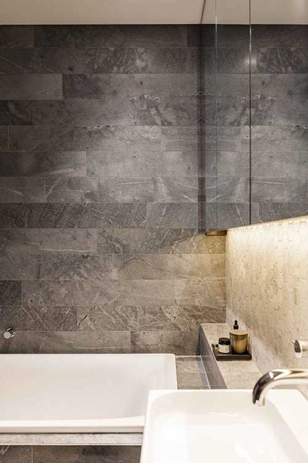 Close-up view of a refined bathroom featuring a large white bathtub, white basin, dark grey stone-look wall and floor tiles, a textured light wall surface, and warm integrated lighting. A small bath-side ledge holds toiletries, while a frameless glass shower panel is visible at the right.