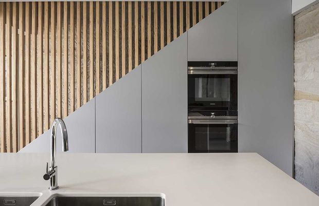 Contemporary minimalist kitchen with a large white stone-look benchtop, integrated stainless-steel sink and tall curved chrome tap. Matte grey handleless cabinetry is arranged beneath a striking diagonal transition to a vertical timber-slat wall, with stacked black built-in ovens and a pale textured masonry wall adding contrast.