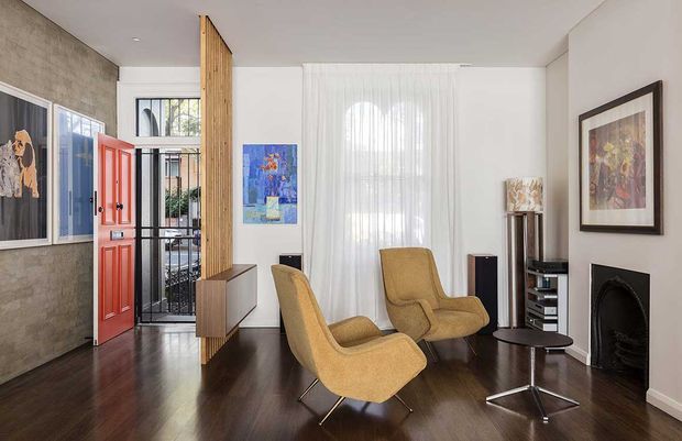 Refined living room with a mid-century-inspired seating arrangement, dark timber flooring, white walls, sheer curtains, framed artwork, a fireplace, and a bright red entry door. A timber screen and glazing define the entry area, while mustard upholstered lounge chairs and a small round coffee table create a warm focal point.