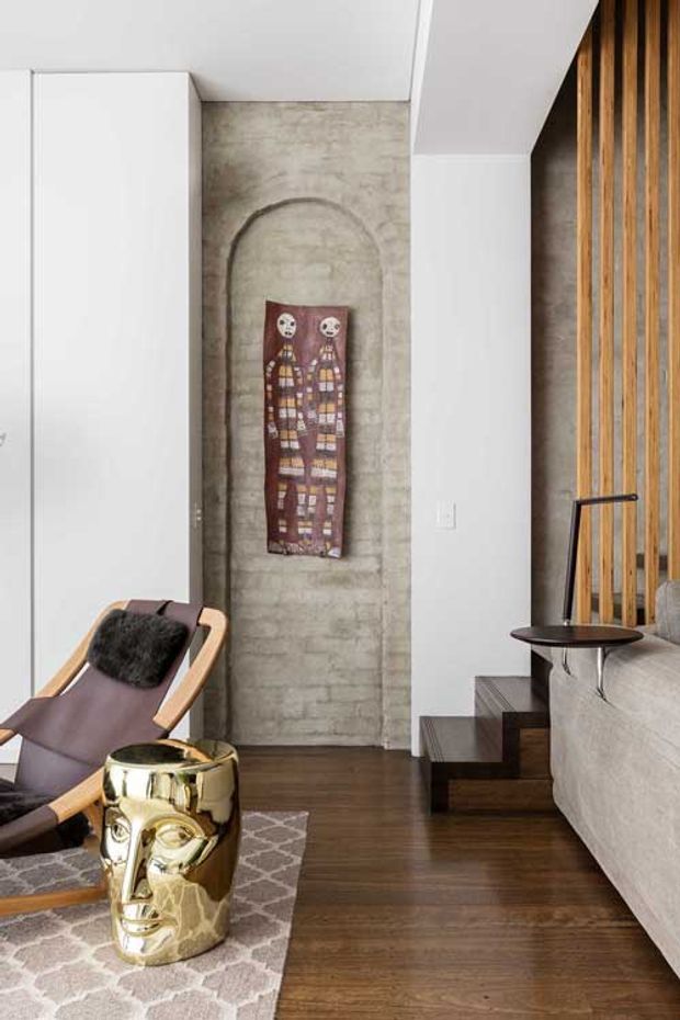 A refined contemporary living room featuring dark timber flooring, a raw concrete feature wall with an arched recessed detail, white built-in cabinetry, vertical timber slat screening, and a compact staircase. The room includes a leather-and-wood lounge chair, sculptural metallic side table, patterned rug, and a built-in light grey upholstered sofa.