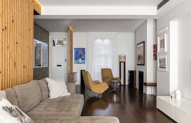 A bright, refined living room with a large neutral sectional sofa, two mid-century-style lounge chairs, dark timber flooring, and a white built-in media wall. Vertical timber battens form a feature wall at left, while a dark textured wall surrounds the entry. Sheer curtains, framed artwork, display shelving, and a linear ceiling light add warmth and detail.
