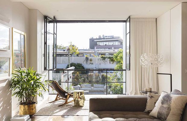 Bright contemporary living room with a large open folding window wall overlooking an urban residential setting. The room features light neutral walls, floor-to-ceiling glazing, sheer curtains, a low upholstered sofa, sculptural lounge chair, indoor plant, and a decorative floor lamp. Natural light creates an airy, refined atmosphere.