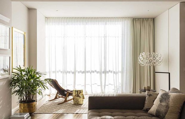 Bright, refined living room with floor-to-ceiling sheer curtains, a neutral sectional sofa, sculptural floor lamp, indoor plant, framed artwork, and a timber rocking chair in a sunlit glazed sitting area. The space has a warm neutral palette and a clean contemporary feel.