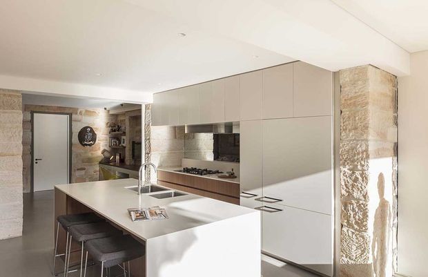 Contemporary open-plan kitchen with a large white island, integrated sink and brushed metal tap, dark upholstered bar stools, and streamlined matte cabinetry in warm grey and timber tones. Exposed rough stone walls and structural columns add rustic texture to the otherwise refined, minimalist fit-out. A dining or living area is partially visible beyond the kitchen.