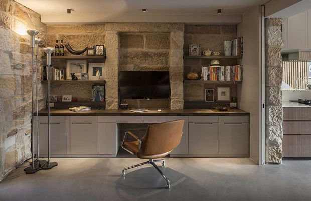 A refined built-in living room media and study wall with low grey cabinetry, open shelving, a central television, books and framed objects. Rough exposed stone walls form a warm textured backdrop, while a sculptural brown leather swivel chair sits on a smooth grey floor. A glazed opening at right reveals part of an adjacent kitchen or utility space.