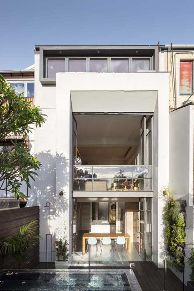 A narrow multi-storey urban townhouse extension viewed from the rear courtyard. The facade is predominantly white rendered masonry with extensive floor-to-ceiling glazing, projecting box-like forms, and a glazed balcony overlooking a small tiled plunge pool. An outdoor dining area is visible on the lower level, with vertical garden planting along the side wall and dark timber screening.