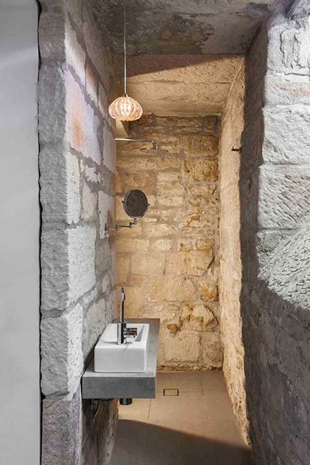 A narrow, rustic bathroom carved into exposed stone masonry, with a wall-mounted white basin on a dark floating vanity, chrome tapware, a round shower fixture, concealed floor drain, and a woven pendant light. The rough natural stone walls and ceiling dominate the space, creating a warm, atmospheric fit-out.