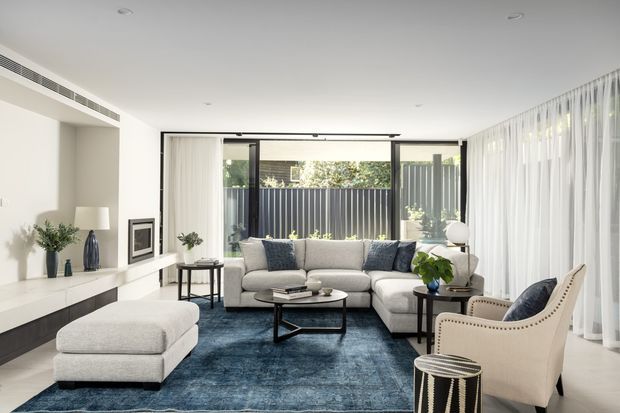 A bright, contemporary living room with a large light sectional sofa, upholstered armchair, dark blue area rug, and black-framed coffee table. Floor-to-ceiling sheer curtains line the glazed wall, opening to a private courtyard or exterior garden, while a long built-in media console runs along the left wall. The palette is predominantly white, light grey, and deep blue with restrained decorative styling.
