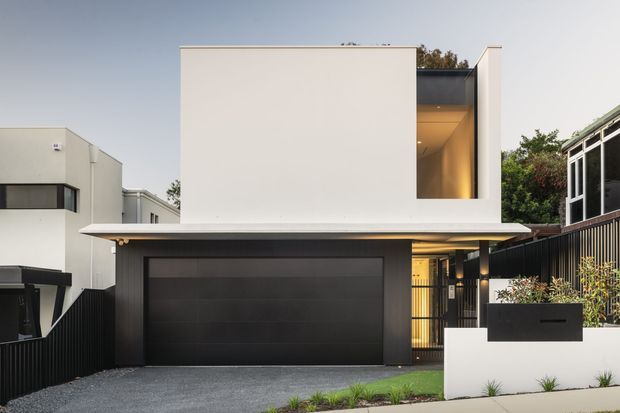 Contemporary two-storey residential facade viewed from the street, featuring a crisp white upper volume, dark charcoal garage door and entry elements, a broad flat canopy, and a recessed warmly lit upper-level opening. A low white rendered boundary wall, black vertical fencing, driveway, and restrained planting complete the frontage.