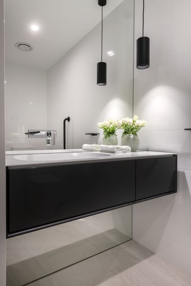 Contemporary bathroom vanity with a wall-mounted glossy black floating cabinet, white integrated basin, pale large-format tiled walls and floor, and a large mirror. Black cylindrical pendant lights hang above the vanity, while a freestanding bath and chrome fittings are visible in the background. White flowers and folded towels add a soft decorative touch.