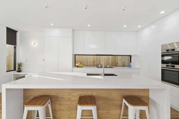 Bright, minimalist white kitchen with extensive flat-panel cabinetry, a large white island with integrated seating, timber-fronted breakfast bar, stainless-steel sink and tap, and built-in black glass ovens. A timber-look splashback adds warmth, while three white stools with timber seats line the island. The room has a clean contemporary appearance and recessed ceiling lighting.