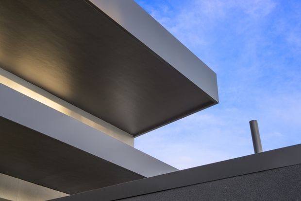 Close-up upward view of a modern building exterior showing overlapping flat roof slabs and projecting box-like forms, with dark textured soffits, smooth light-grey fascia bands, and a cylindrical metal flue against a blue sky. The image emphasizes strong horizontal planes and minimalist geometric composition.