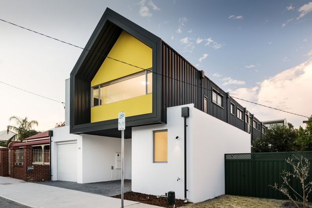 Contemporary two-storey urban residence with a bold black-framed projecting gable volume, bright yellow feature wall and clerestory glazing above a crisp white ground-floor facade. Vertical dark cladding extends along the upper level, with an integrated garage, boundary fencing and neighbouring homes visible at street level.
