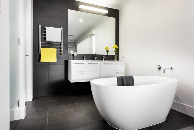 Contemporary bathroom featuring a freestanding white bathtub, wall-mounted white vanity with integrated storage, large rectangular mirror, dark charcoal wall and floor tiles, and a chrome heated towel rail with white and yellow towels. The room has bright white walls, recessed ceiling detailing, and simple modern fixtures.