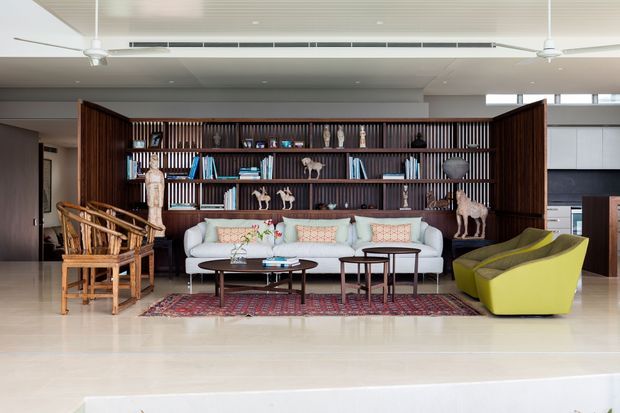 A bright, open-plan living room centred on a large dark timber display bookcase with vertical slatted detailing. The room includes a pale sofa, woven timber armchairs, sculptural lime-green lounge chairs, nesting coffee tables, a patterned rug, decorative objects and books. Recessed ceiling lighting and ceiling fans contribute to a spacious, refined contemporary interior.