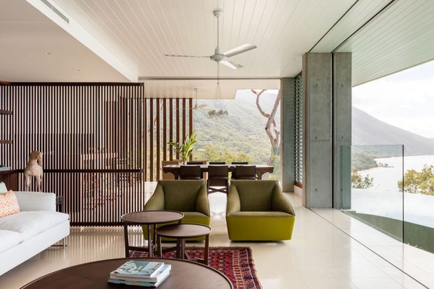 A bright, open-plan living room with a white sofa, olive-green lounge chairs, round coffee tables, a dining area, and a patterned rug. Floor-to-ceiling glazing opens to a balcony or terrace with expansive mountain and water views. The room features a white-lined ceiling, polished light flooring, concrete structural columns, and a dark vertical timber slat screen separating the living and dining zones.