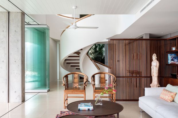 Bright contemporary living room with a sculptural curved staircase, open-riser timber treads, a ceiling fan, polished light flooring, and a large vertical timber slat screen concealing built-in storage or a media area. The room includes a light sofa, timber-and-cane lounge chairs, a round dark coffee table, a tall green-tinted glass feature enclosure, and high ceilings with a clean white palette.