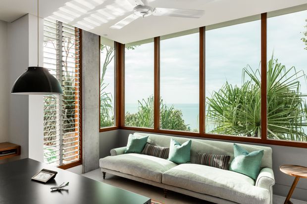 Bright contemporary living room with a large upholstered sofa, green and striped cushions, and expansive timber-framed windows overlooking lush vegetation. A black pendant light hangs over a dark surface in the foreground, with white louvred shutters, a ceiling fan, and strong striped sunlight creating a relaxed indoor-outdoor feel.