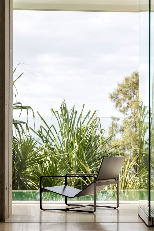 A minimalist interior framed by expansive floor-to-ceiling glazing, with a modern black-framed lounge chair positioned on a pale tiled floor. Dense tropical planting and a narrow turquoise pool or water feature are visible immediately outside, creating a strong indoor–outdoor connection. The space is bright, sparse, and contemporary, with light timber-toned window framing and clear glass.
