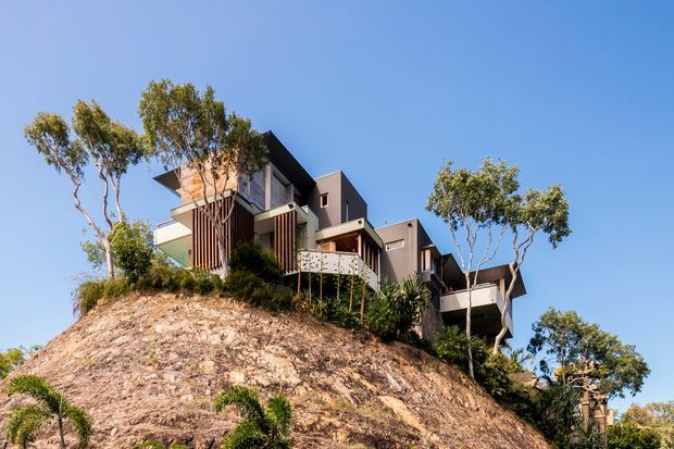 A contemporary multi-level residence stepping down a steep, landscaped hillside. The building has dark charcoal cladding, projecting box-like volumes, extensive glazing, balconies, and vertical timber screening, surrounded by mature trees and planting beneath a clear blue sky.