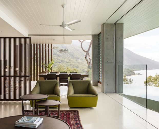 A bright, open-plan contemporary interior with a living area in the foreground and a dining area beyond. Olive-green lounge chairs, round dark occasional tables, a patterned rug, and a dining table with upholstered chairs sit beside full-height glazing overlooking a mountainous, lakeside landscape. Vertical timber screening, a white-lined ceiling with ceiling fan, polished light flooring, and a concrete structural column define the space. A glass balustrade suggests an elevated terrace or balcony beyond the glazing.