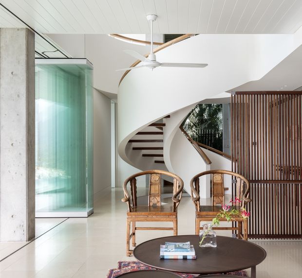 Bright contemporary interior featuring a sculptural curved staircase with dark timber treads, a white painted balustrade, a large ceiling fan, polished light flooring, and a vertical timber screen dividing the space. Two carved timber armchairs and a round dark coffee table occupy the foreground. A translucent green glass wall and exposed concrete column add contrasting materials.