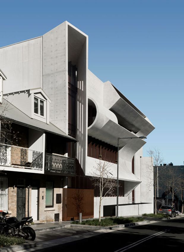 Street-level exterior view of a contemporary multi-storey urban building with a sculptural pale concrete facade, deep geometric projections, curved openings, vertical slit windows, and dark recessed glazing. It sits directly beside an older residential building with ornate balcony railings, creating a strong contrast between modern and heritage architecture. A paved street, sidewalk, young street trees, and parked vehicles are visible.