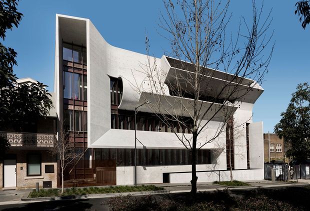 Street-facing view of a distinctive multi-storey contemporary building with sculptural curved white rendered walls, projecting cantilevered upper forms, extensive dark-framed glazing, and a tall vertical glazed entrance or stair volume. The building sits in an urban residential streetscape with mature street trees and neighbouring houses.