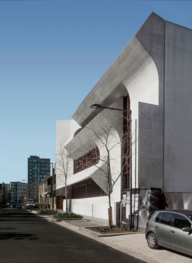 Street-level view of a striking urban concrete building with sculptural, cantilevered and curved facade elements, deep arched openings, vertical metal screens, and a largely blank side wall. The building sits on a city street with neighbouring mid-rise buildings and parked cars visible in the background.